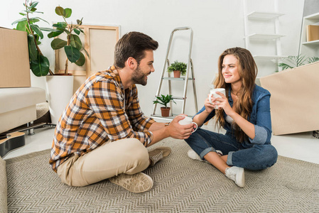 微笑夫妇与杯子咖啡坐在地板在新的房子, 移动的家庭概念