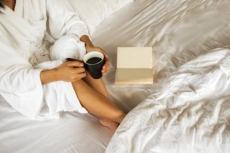 美丽的亚洲女性，坐在床上，一杯咖啡，读一本书。一本书和一杯咖啡的早晨。放松的概念。复古的滤镜效果 柔焦 选择性焦点