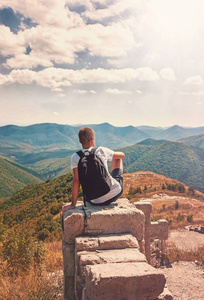 大山太阳他背对摄影师的大石头上坐着,看着大山,天空和云彩的视图上