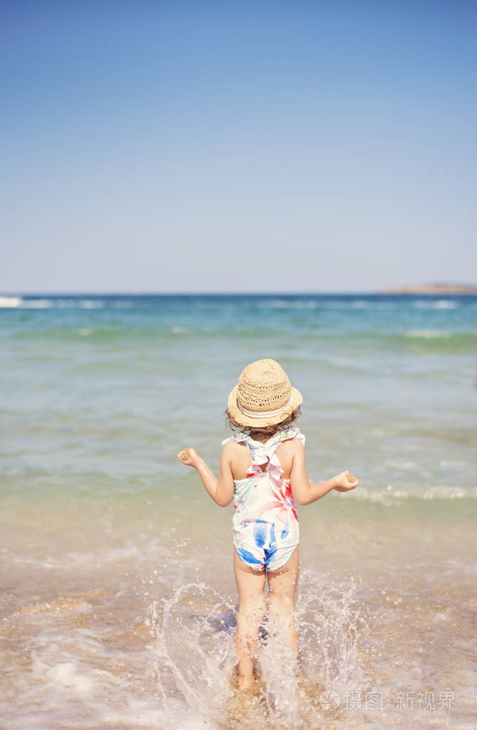 小女孩,孩子与草帽和花卉泳装看着大海,在晴朗的一天