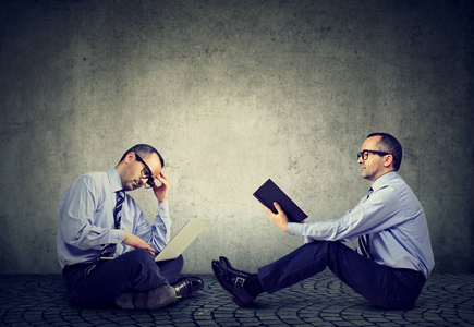 两名男子坐在地上一读一本书在笔记本电脑上的另一个工作