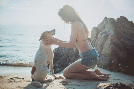 肖像的美丽快乐的年轻女子坐在海滩玩她的宠物狗日落海岸黄昏的作为背景