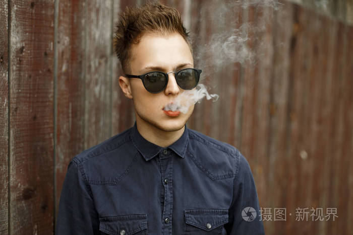 时尚的年轻人,在烟雾弥漫的太阳镜抽木立在墙边的香烟照片-正版商用