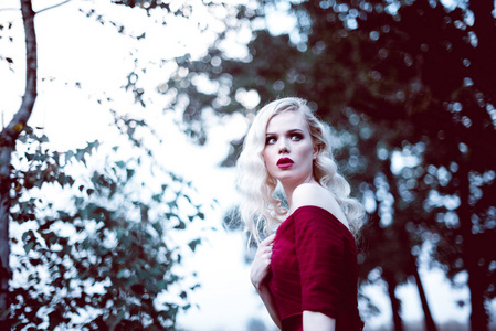 时尚华丽年轻的金发女郎在漂亮的红色裙子，在一个童话般的森林。魔幻的气氛。调色射击