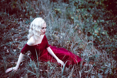 时尚华丽年轻的金发女郎在漂亮的红色裙子，在一个童话般的森林。魔幻的气氛。调色射击