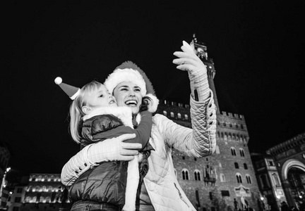 在佛罗伦萨的圣诞节期间充满灵感的旅程。愉快的年轻母亲和儿童旅行者在圣诞节帽子反对宫殿旧在佛罗伦萨, 意大利拥抱和采取自拍