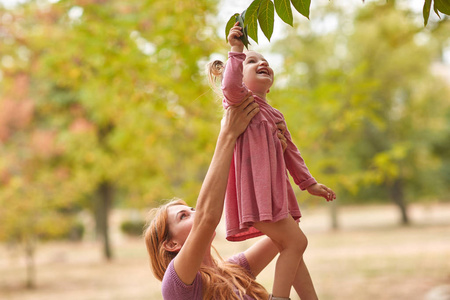 快乐的妈妈带着孩子在公园里走在阳光灿烂的日子 触摸和考虑上一棵树上的叶子