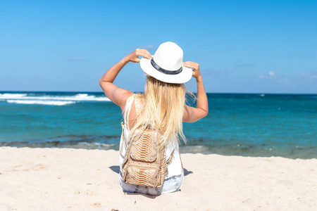 年轻的女人，与白色的帽子和豪华蛇皮 python 背包坐在白色的沙滩上一个热带的巴厘岛在阳光明媚的日子。海洋成本