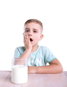 在蓝色 t 恤的小可爱男孩非常吃惊，坐在旁边充满健康的牛奶站在玻璃上的棕色的桌子，白色背景上孤立