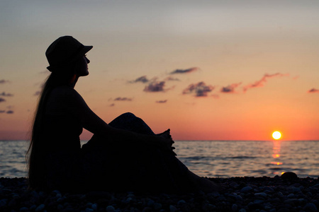 女孩在日落时分的大海背景上坐一顶帽子。剪影