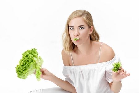 美丽的金发碧眼女人吃的新鲜绿色沙拉在白色背景上的白色衬衫。健康与饮食