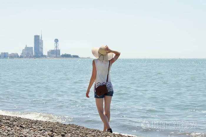 戴帽子的女孩沿着海边散步。在距离城市。查看从背面