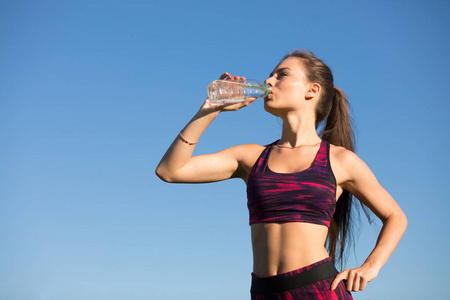 年轻快乐的女人从瓶里喝水。在运动服在蓝色天空背景在户外举行喝的孤立的运动员。她止渴