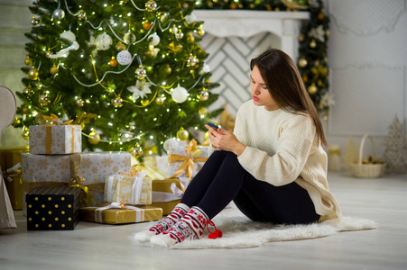 可爱的黑头发女孩坐在附近的圣诞树用手机在手中