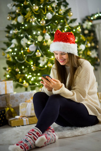 好的女孩，在圣诞老人的帽子坐在附近的圣诞树用手中的手机
