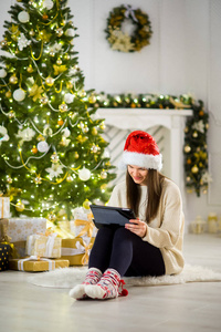迷人的女孩，在圣诞老人的帽子坐在附近的一棵圣诞树与笔记本计算机