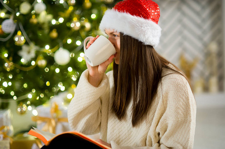在圣诞老人的帽子的女孩喝咖啡附近圣诞树