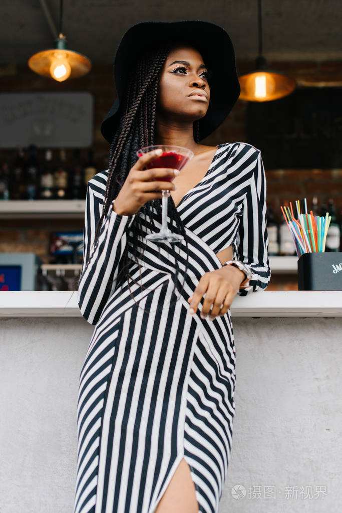 美国女人在酒吧与红色鸡尾酒在她的手中在很长的条纹裙子的漂亮女人