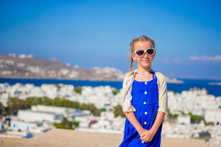 穿着蓝色衣服在户外的小女孩。在街的典型希腊传统村庄与白色的墙壁和多彩门在希腊米克诺斯岛上的孩子