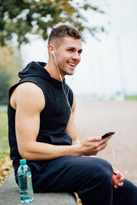 健身男人穿着运动服听听音乐，坐在一张石凳上。在艰苦的锻炼，说话后放松一下