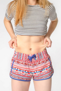 年轻女子腹部肚皮穿着短裤和作物顶部。她拉扯皮肤两边，显示多余的脂肪。健康的饮食和体重损失的概念