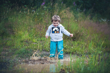 可爱的卷发宝宝穿着靴子和橡胶裤子在水坑里跳。有趣的孩子，与孩子们的幸福