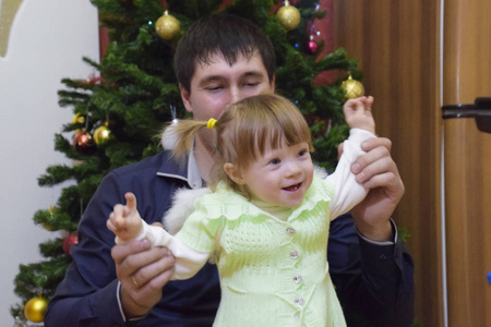 一岁的女孩和爸爸在圣诞树的背景下。 一个有灰色眼睛和金发的孩子。