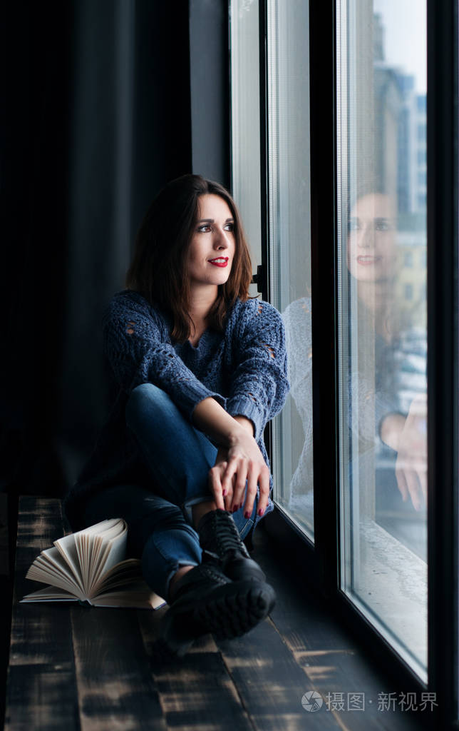 蓝色牛仔裤和黑色棕色头发的美丽年轻女子 (女孩) 皮靴,坐在窗台上