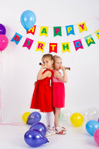 两个女孩正在吹口哨，站在旁边的气球题字生日快乐，白色背景