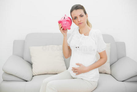 失望的孕妇抱着肚子摇着粉红色的存钱罐