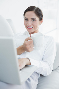 带着笔记本电脑和咖啡杯在沙发上微笑的穿着考究的女人