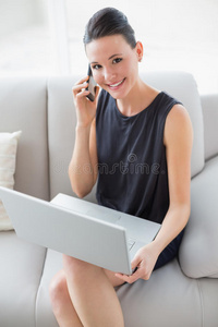 坐在沙发上用手提电脑和手机的漂亮的笑容灿烂的穿着考究的女人