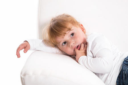 可爱的害羞的女孩躺在白色沙发上吮吸拇指或手指