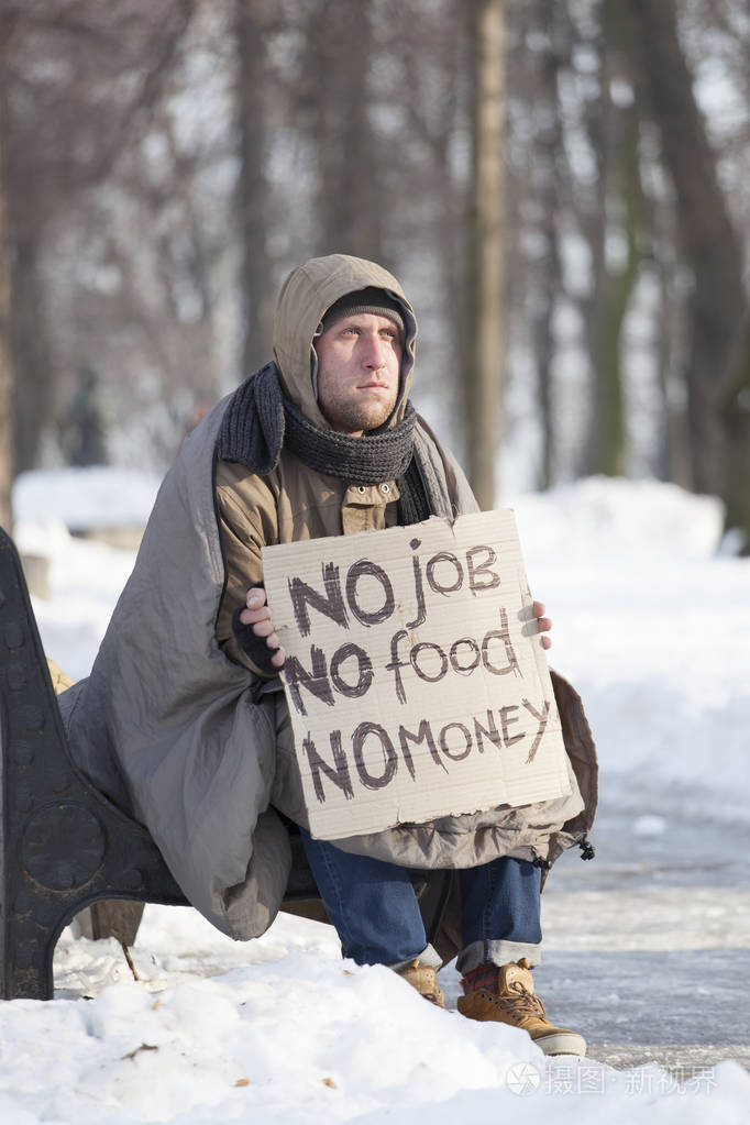 年轻饿的无家可归的人在冬季城市公园的纸板求助