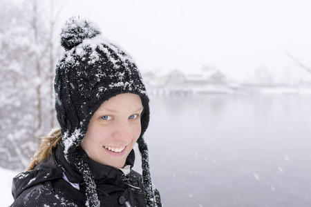 冬季的针织黑色羊毛帽子与雪的蓝眼睛的年轻女子肖像期间降雪片高山湖泊和乡村背景