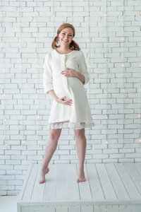 年轻怀孕女孩微笑着，站在白色的墙壁附近