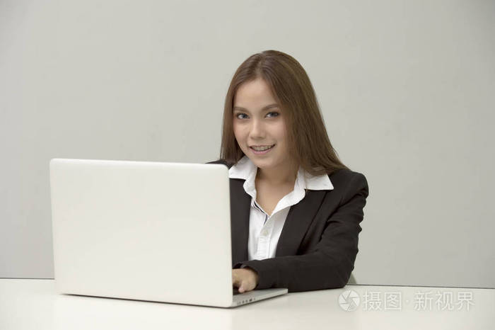 专业女商人，同时看着相机，微笑着坐在她的书桌里的办公笔记本电脑