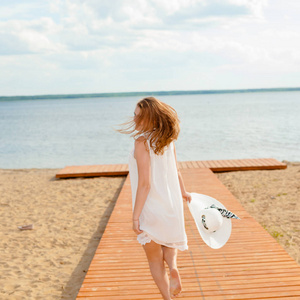 浪漫的女孩，在白色的连衣裙，戴着白色帽子转向长尾的木码头在海滩上