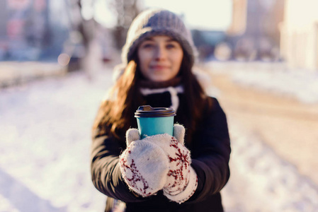 美丽的金发白种人年轻女子在冬天的夹克，针织帽子豆豆，捧着咖啡或茶，在下雪的冬天在户外在公园外卖杯。选择性的焦点