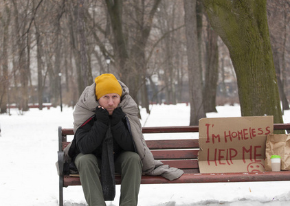 坐在板凳上的乞丐用硬纸板在冬季城市公园成人无家可归的人坐在长凳上