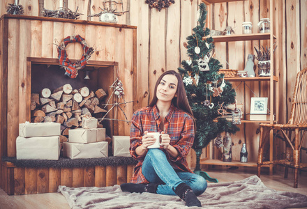 很漂亮，性格开朗的年轻女孩的圣诞树在格子衬衫与在她手中的红色咖啡杯背景上布鲁内特的肖像。新的一年的概念。女孩微笑着，休息