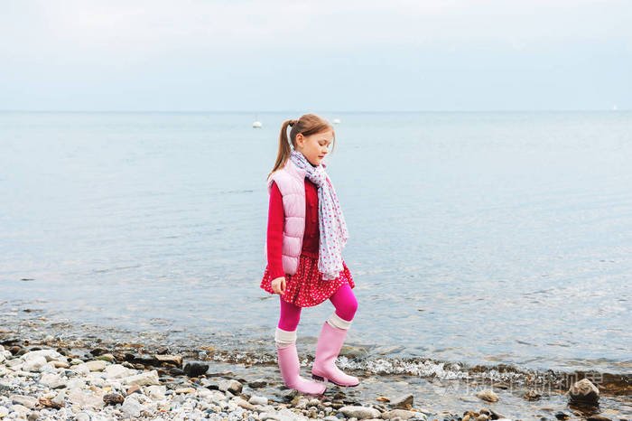 可爱的小女孩在湖边散步在寒冷阴天穿着粉红色的背心和雨靴