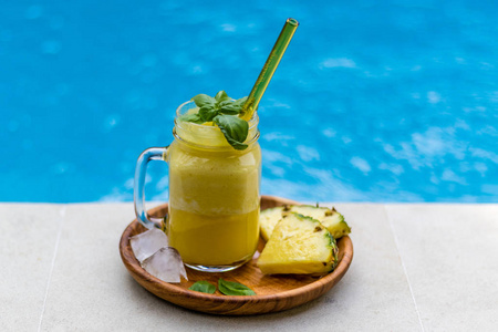 鲜榨菠萝汁用黄色玻璃稻草 黑暗的木制板，玻璃配两片菠萝，泳池的边缘上的冰和罗勒叶的多维数据集。侧视图