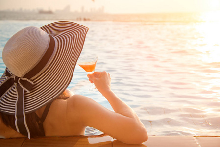 年轻的女人在游泳池边，在夕阳海边放松的大帽子