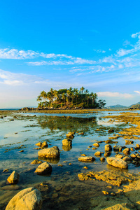 当海水在蓝天和阴天背景下退去时，带着岩石海滩和树木的偏僻小岛