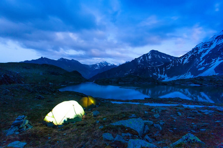 发光的帐篷站在银行的山间溪流，高山峰，白雪皑皑的雪峰。黄昏，夜。一群游客正在休息