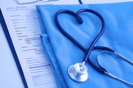 医用听诊器扭曲心形躺在病人医疗历史记录列表和蓝色医生统一特写。医疗帮助或保险的概念。心脏病学护理 保健 保护及预防