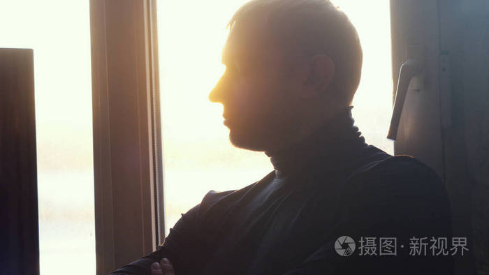 年轻的疲惫的男人坐在窗边, 在日落的时候, 透过太阳看着办公室里肮脏