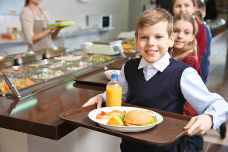 可爱的女孩拿着托盘和美味的食物在学校食堂照片