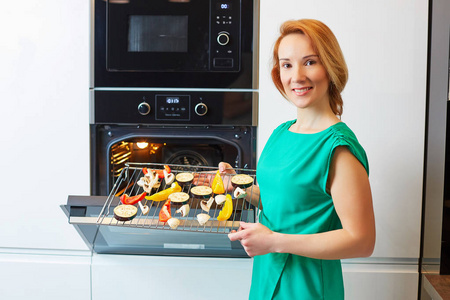 漂亮微笑的女人红头发站在烤箱附近和持有格栅与蔬菜，看着相机在现代厨房